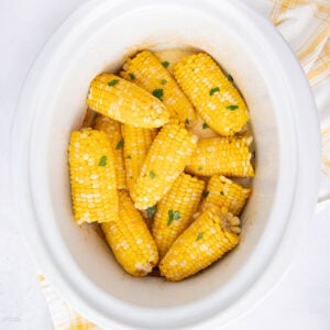 Corn on the cob pieces garnished with chopped parsley inside a white slow cooker, with a yellow and white striped cloth beside it.