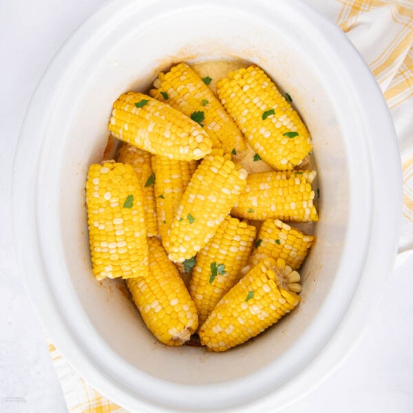 Corn on the cob pieces garnished with chopped parsley inside a white slow cooker, with a yellow and white striped cloth beside it.