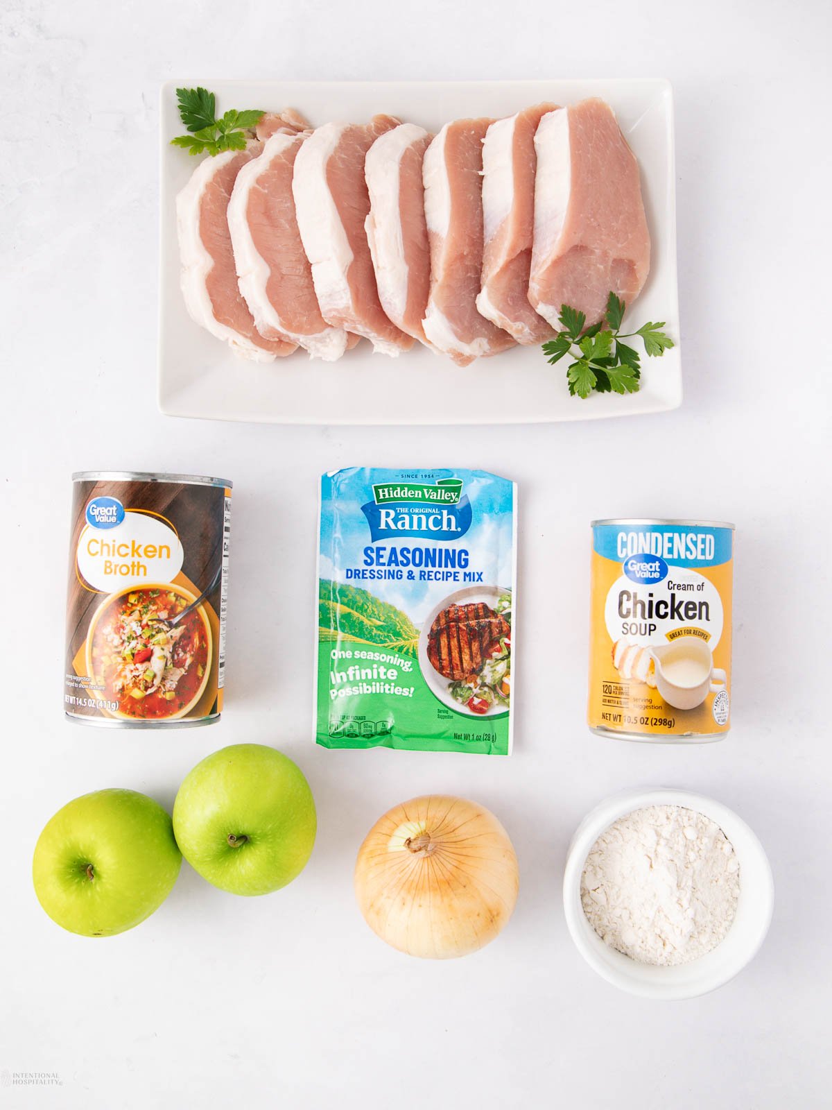 Raw pork chops on a rectangular white plate with parsley, surrounded by cans of chicken broth and cream of chicken soup, a packet of ranch seasoning, two green apples, a yellow onion, and a bowl of flour.