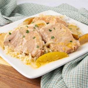 A square white plate holds cooked pork chops with onions and apple slices served over rice, garnished with chopped herbs. A blue patterned cloth is underneath the plate on a wooden surface.