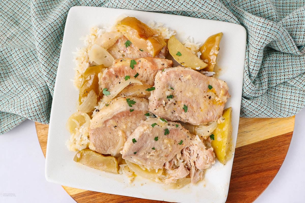 A square white plate with slices of cooked pork, onions, apple wedges, and a light sauce, served over rice. The dish is garnished with chopped herbs, and a checkered cloth napkin is in the background.