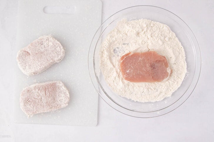 Raw pork chops being coated in flour; one pork chop is in a glass bowl of flour, while two floured pork chops are resting on a white cutting board, all on a white surface.