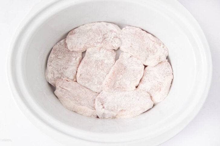 Raw, flour-dusted pork chops arranged in a single layer inside a white slow cooker, ready for cooking.