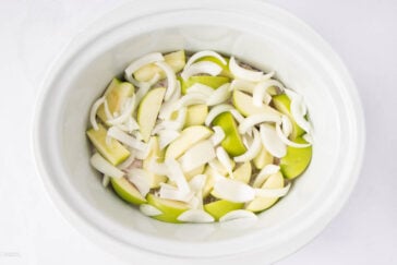 A white slow cooker containing sliced green apples and raw onion slices, arranged in an overlapping layer.