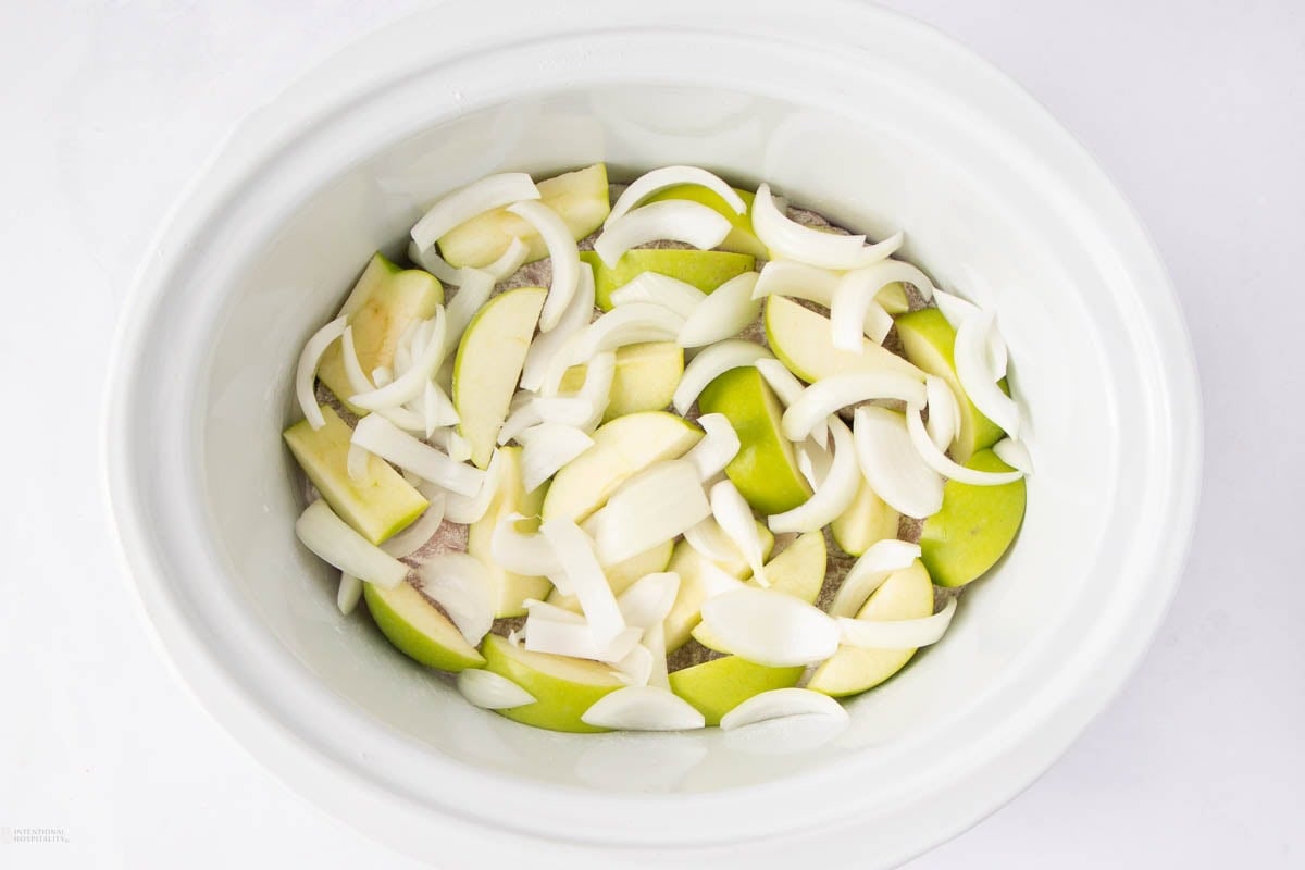 A white slow cooker containing sliced green apples and raw onion slices, arranged in an overlapping layer.