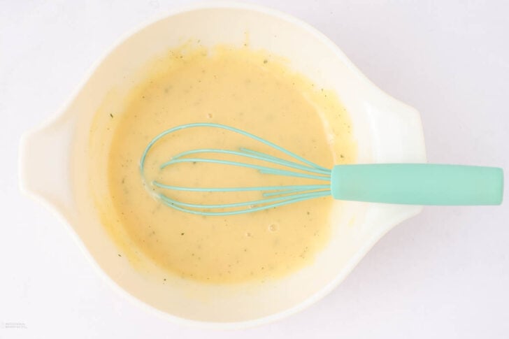 A white mixing bowl containing a pale yellow batter being whisked with a light blue whisk on a white surface.