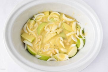 Sliced green apples and onions covered in a creamy yellow sauce inside a white slow cooker, ready for cooking.