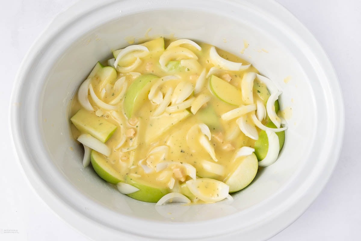 A white slow cooker filled with cooked sliced potatoes, chunks of sausage, and creamy sauce, sitting on a light checkered cloth.