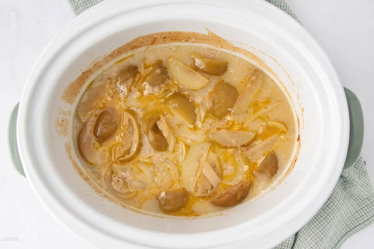 A white slow cooker filled with cooked sliced potatoes, chunks of sausage, and creamy sauce, sitting on a light checkered cloth.