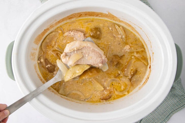 A hand holds a spatula lifting a piece of cooked chicken with onions and sauce from a white slow cooker. The slow cooker contains more chicken, onions, and creamy sauce. A green kitchen towel is partially visible underneath.