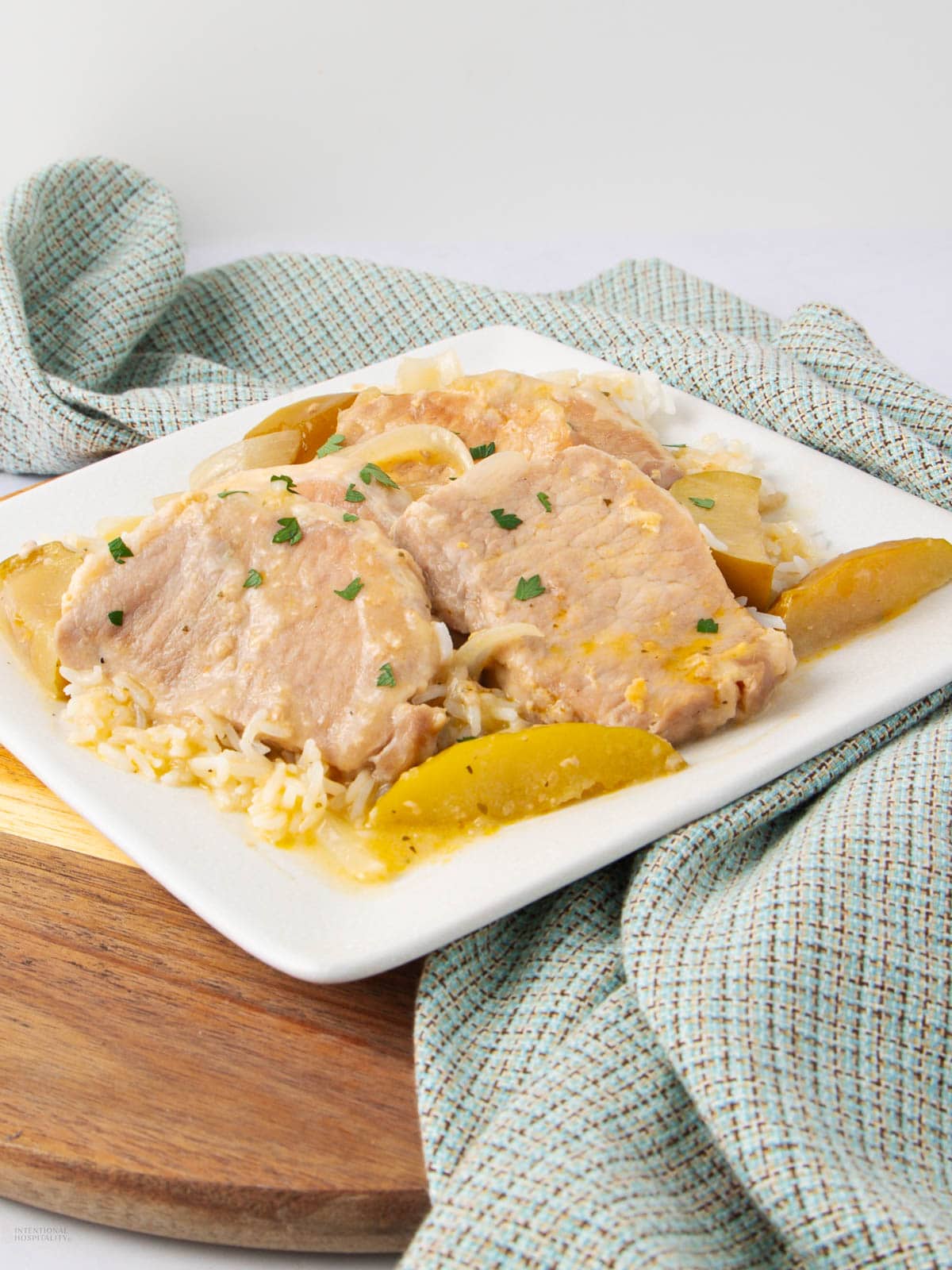 Three slices of pork with cooked apple wedges and onions served over rice on a white square plate, garnished with chopped herbs. A blue checkered cloth and wooden board are in the background.
