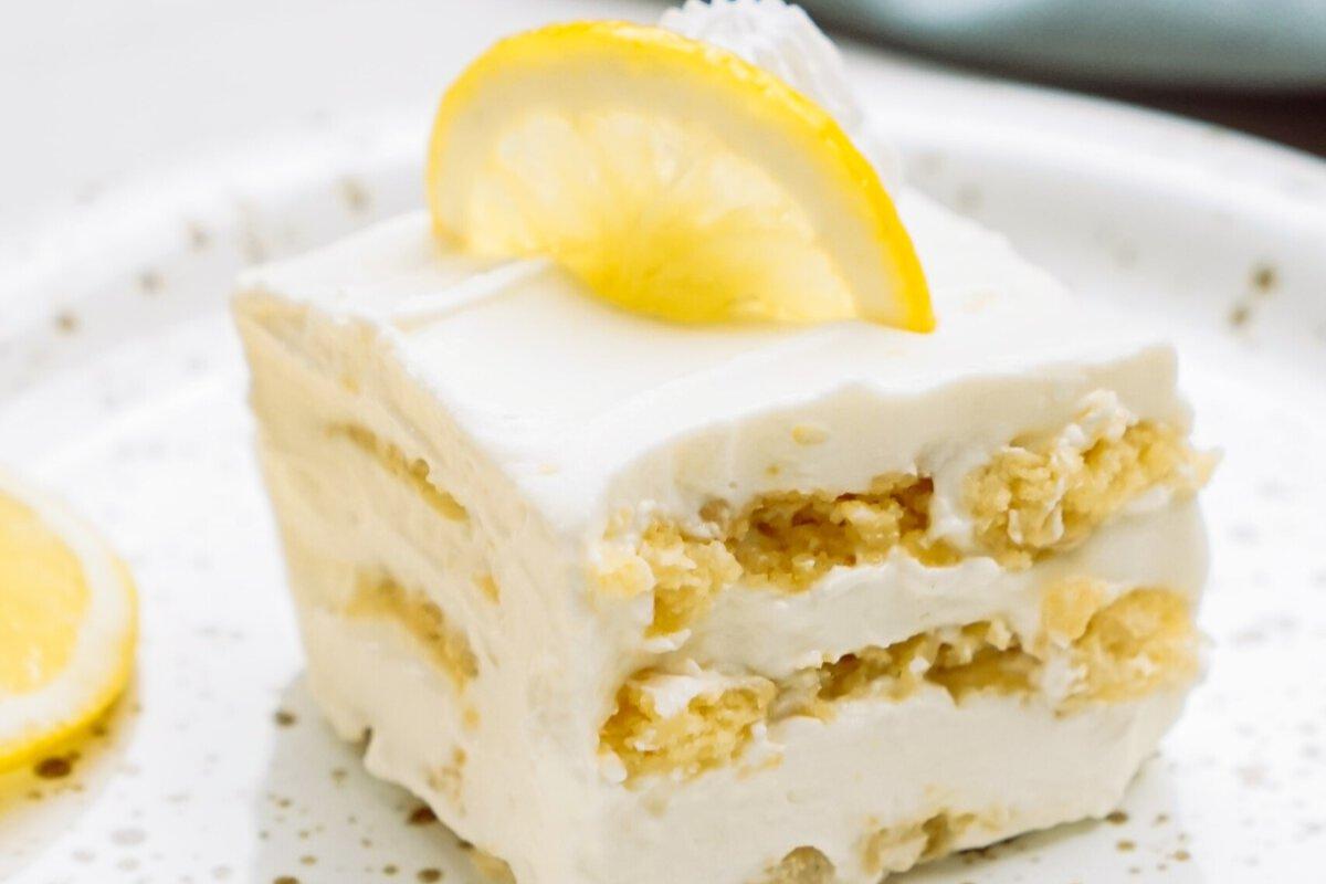 A slice of creamy lemon dessert sits on a white plate, featuring layers of fluffy filling and cake, and topped with a fresh lemon slice.