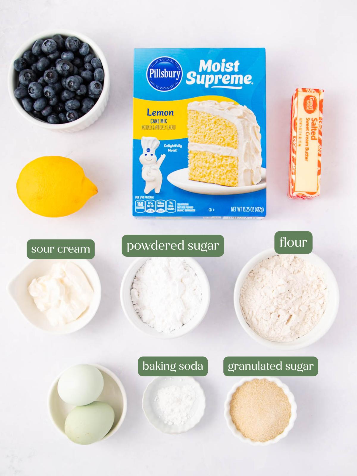 Top-down view of ingredients for a lemon blueberry dessert: a box of lemon cake mix, butter, blueberries, a lemon, sour cream, powdered sugar, flour, two eggs, baking soda, and granulated sugar in bowls. Labels indicate some items.