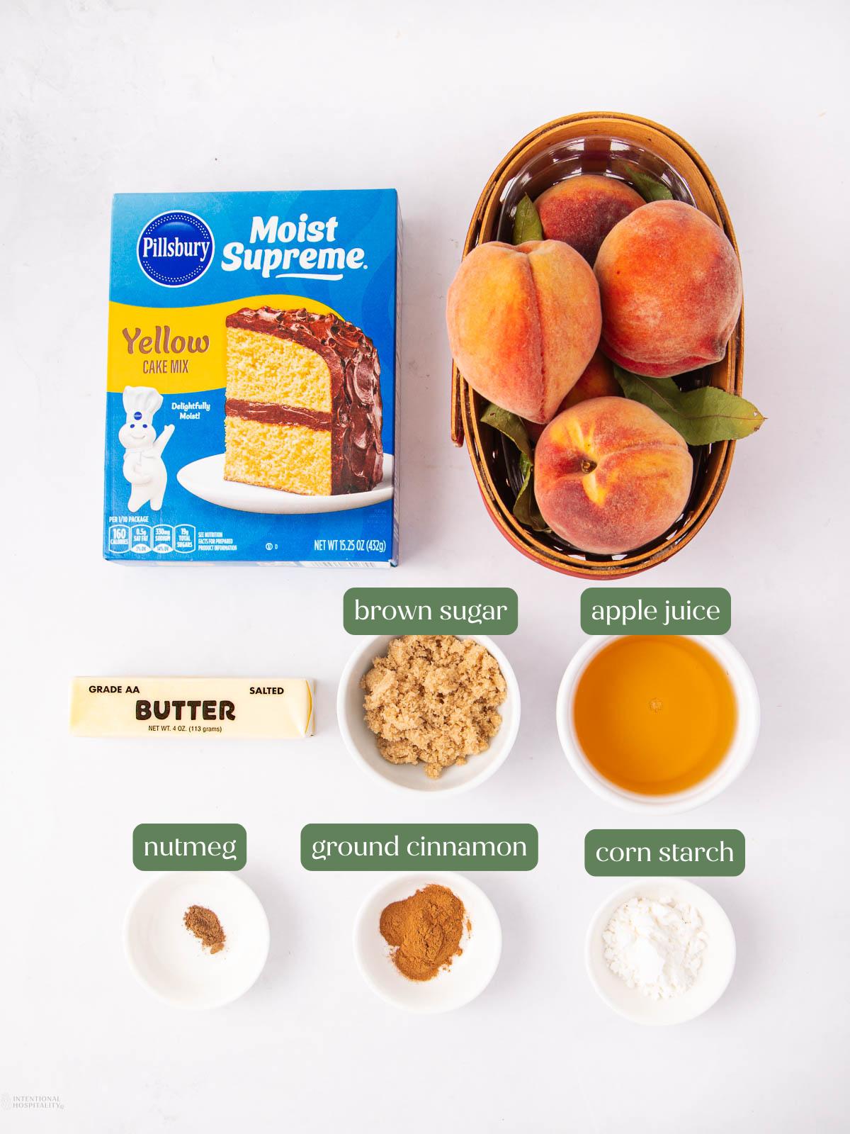 Ingredients for a peach dessert displayed on a white background: yellow cake mix, fresh peaches in a basket, brown sugar, apple juice, a stick of butter, nutmeg, ground cinnamon, and corn starch in small bowls.