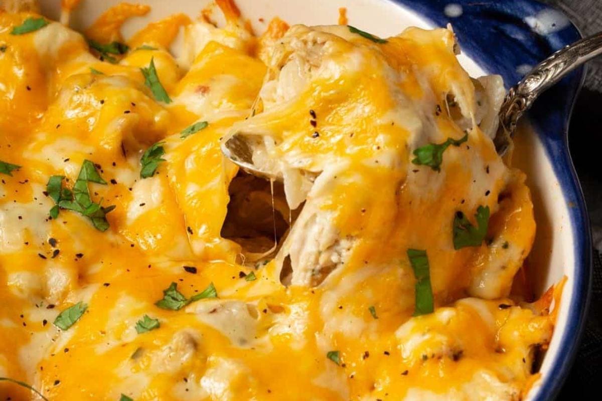 A close-up of a cheesy chicken casserole topped with melted cheddar and mozzarella cheese, garnished with chopped parsley, with a spoon lifting a serving.