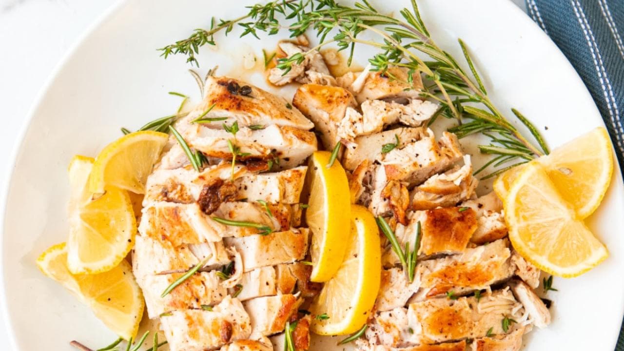 Sliced grilled chicken breast garnished with fresh rosemary and lemon wedges on a white plate, with a blue and white napkin partially visible at the edge.