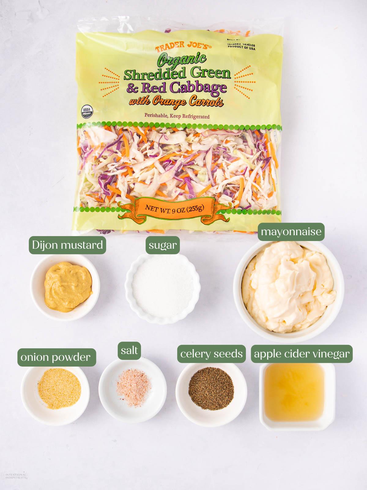 A bag of Trader Joe’s shredded green and red cabbage with carrots is on a white surface, surrounded by small bowls containing mayonnaise, Dijon mustard, sugar, salt, celery seeds, onion powder, and apple cider vinegar.