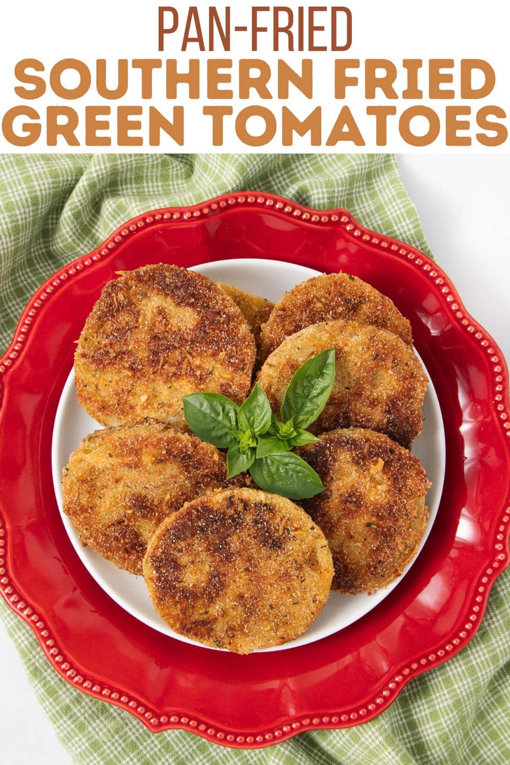 A white plate with six golden-brown fried green tomato slices is placed on a red decorative plate. The tomatoes are garnished with fresh basil and set on a green checkered cloth. Text above reads "Pan-Fried Southern Fried Green Tomatoes.