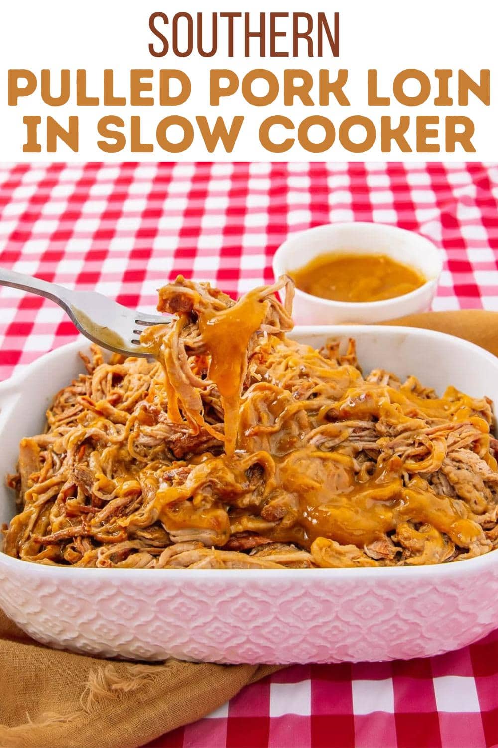 A white dish of shredded pulled pork loin topped with sauce sits on a red and white checkered tablecloth. A fork lifts a portion, and a small bowl of sauce is in the background. Text above reads, "Southern Pulled Pork Loin in Slow Cooker.