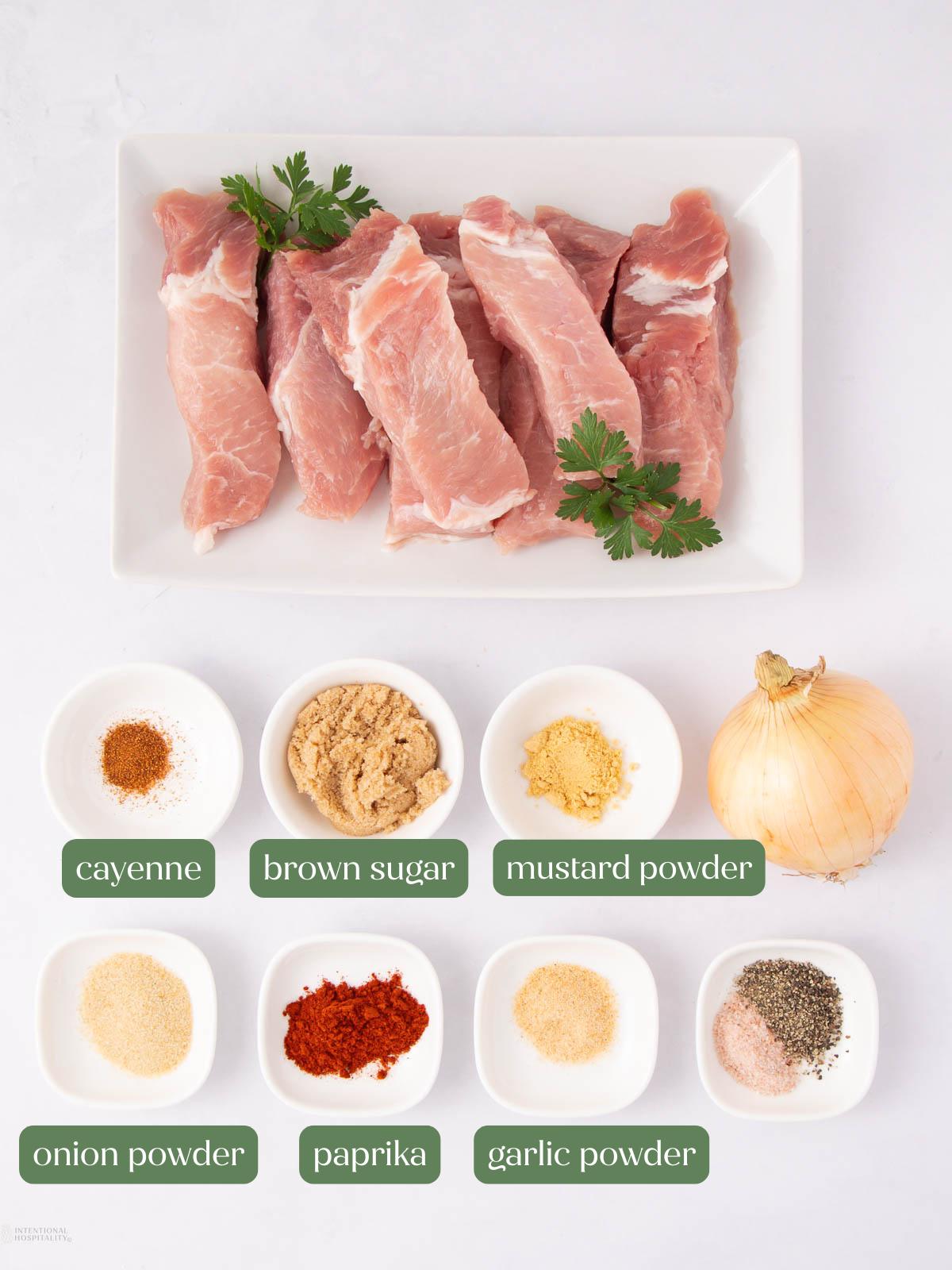 Raw pork ribs on a white plate garnished with parsley, surrounded by small bowls holding cayenne, brown sugar, mustard powder, onion powder, paprika, garlic powder, black pepper, and a whole onion.