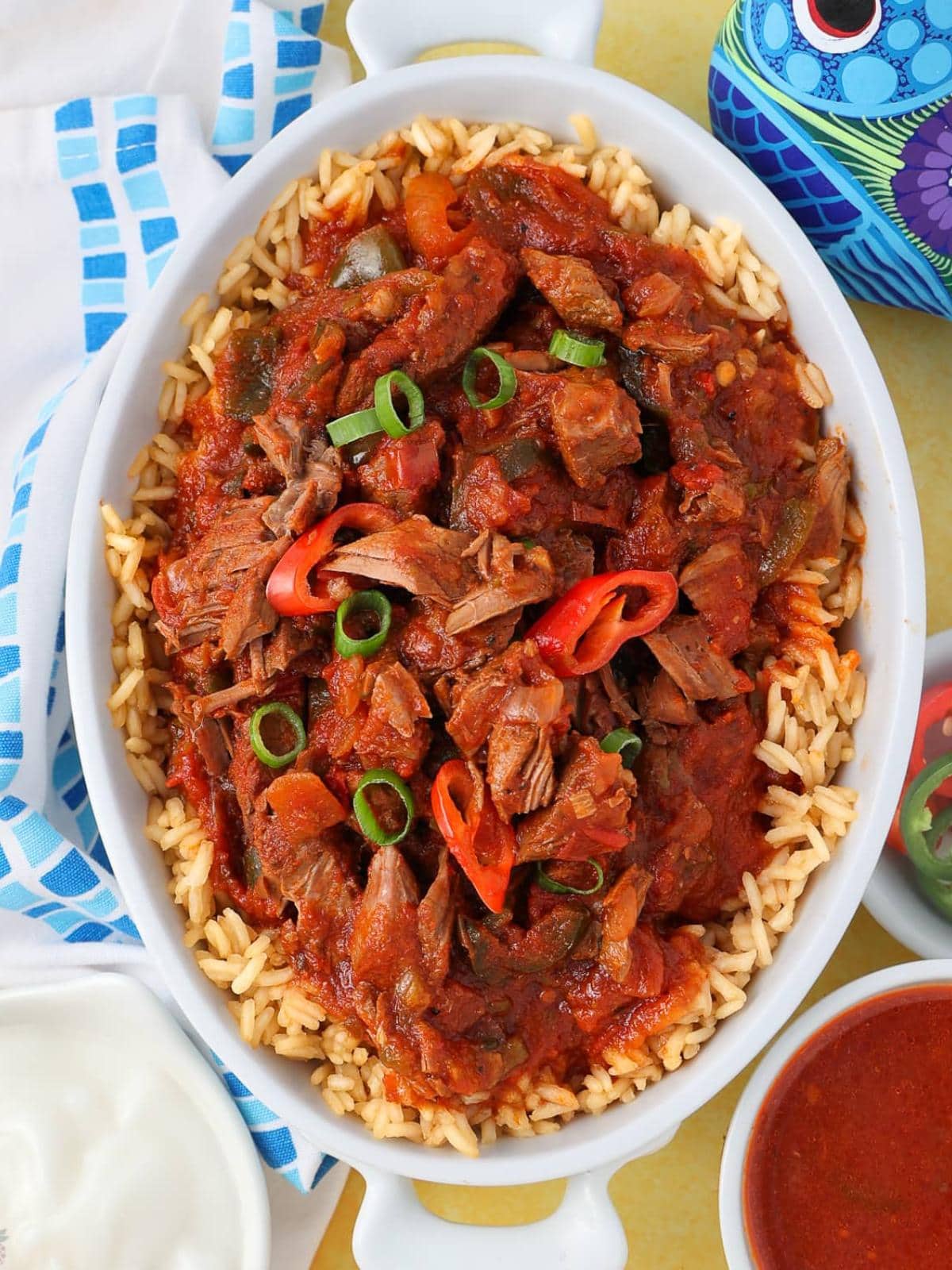 A white oval dish filled with rice topped with shredded beef in a rich red tomato sauce, garnished with sliced green onions and red chili peppers. Surrounding the dish are a blue patterned jug, a napkin, and bowls of sauce and sour cream.