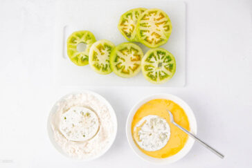 Sliced green tomatoes on a cutting board, with one slice coated in flour in a bowl and another slice being dipped in beaten eggs in a separate bowl, ready for breading and frying.
