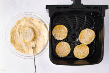 A bowl of breaded slices with a spoon beside an air fryer basket containing four evenly spaced breaded slices, ready to be cooked. The background is a clean white surface.
