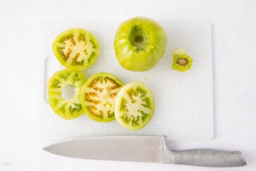 A whole green tomato, several sliced green tomatoes, a tomato stem, and a large knife are placed on a white cutting board against a white background.