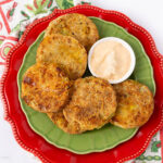 A green plate holds several fried green tomato slices with a small bowl of creamy dipping sauce. The plate is set on a larger red plate, with a colorful patterned cloth nearby.