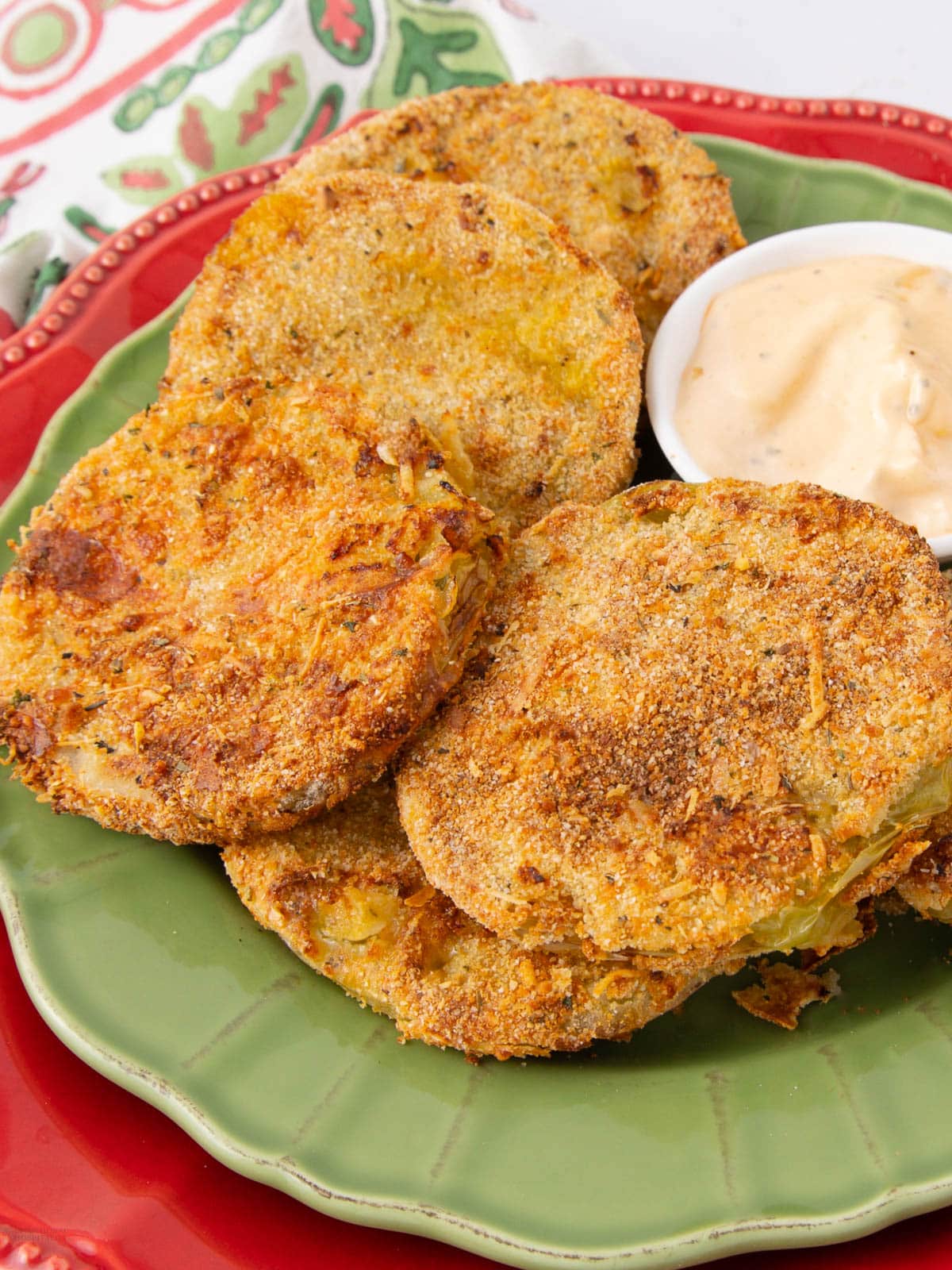 A green plate with several golden-brown fried green tomato slices, served with a small dish of creamy dipping sauce, sits on a red charger with a colorful napkin in the background.