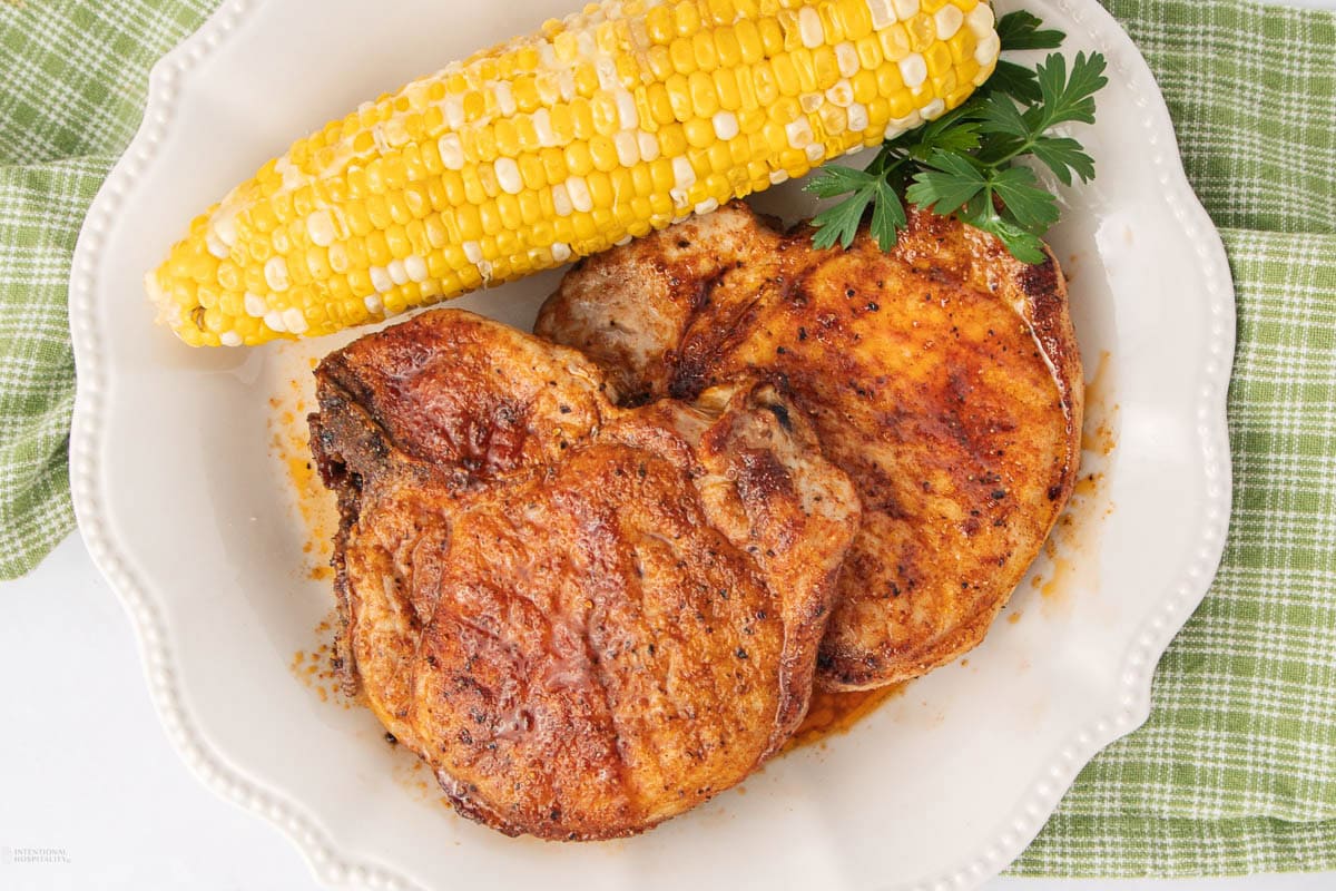 Two seasoned, cooked pork chops and an ear of corn on the cob are served on a white plate, garnished with a sprig of parsley, atop a green checkered cloth.