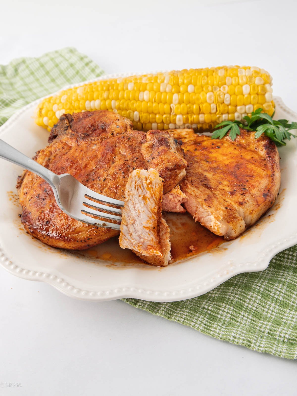 A plate with two seasoned pork chops, one sliced with a fork, served with a corn on the cob and garnished with parsley, placed on a green checkered cloth.