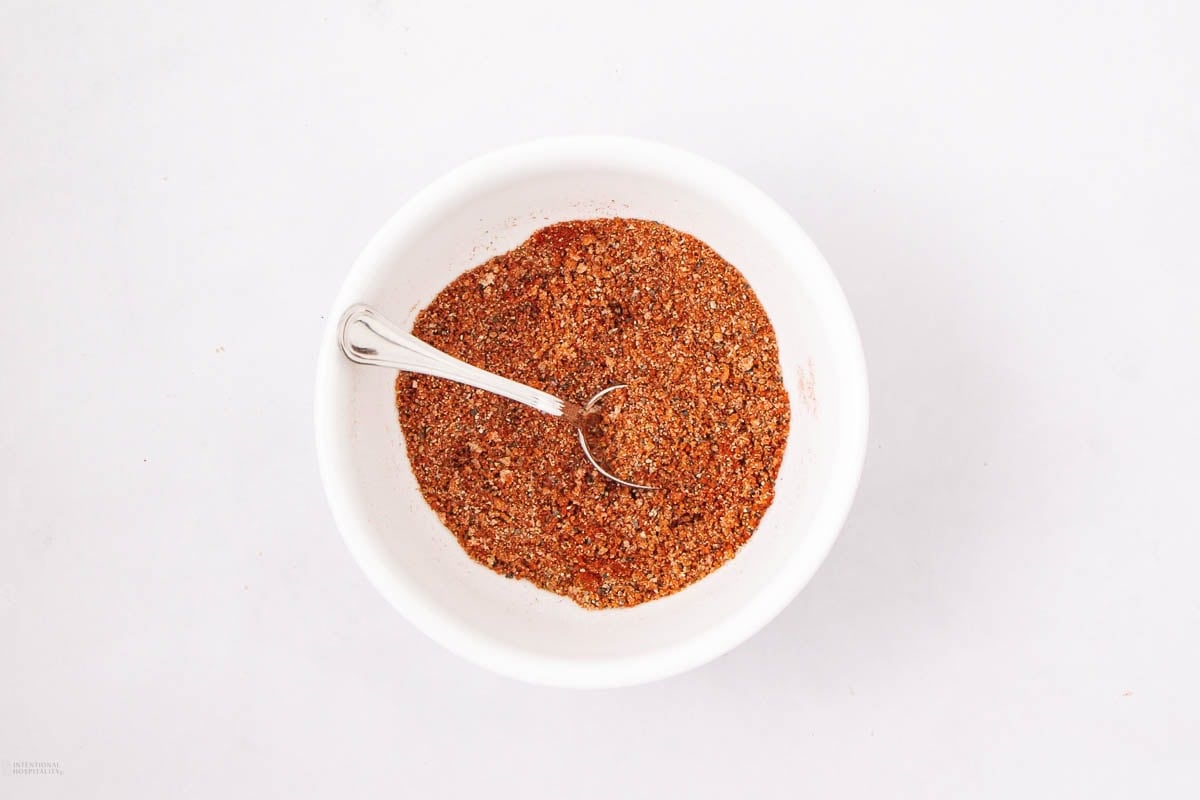 A white bowl filled with a reddish-brown spice mix, with a metal spoon resting inside, on a plain white background.