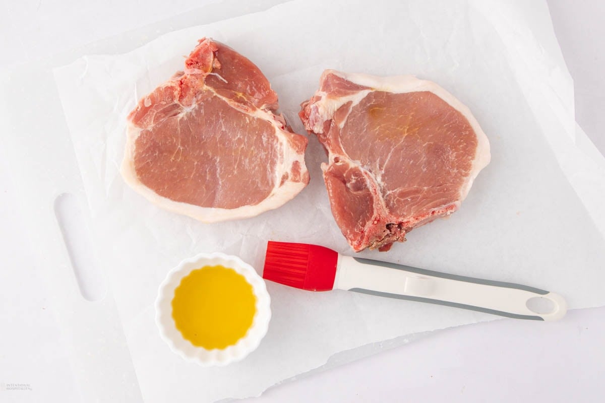 Two raw pork chops on white parchment paper next to a small white bowl of olive oil and a red silicone basting brush with a white handle, all placed on a white surface.