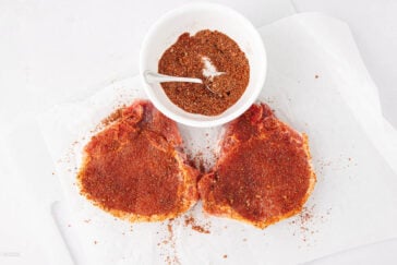 Two raw pork chops covered in a reddish-brown spice rub sit on white parchment paper next to a small white bowl filled with more seasoning and a spoon.