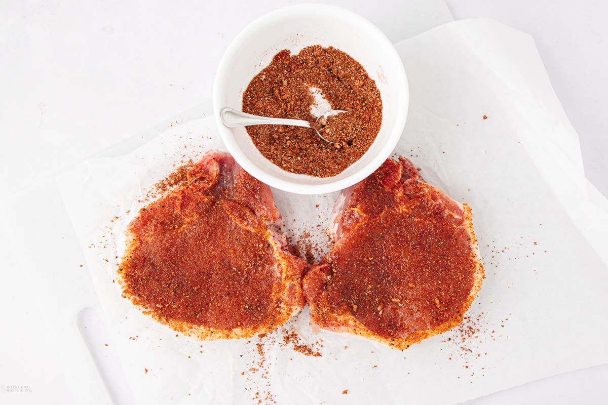 Two raw pork chops covered in a reddish-brown spice rub sit on white parchment paper next to a small white bowl filled with more seasoning and a spoon.