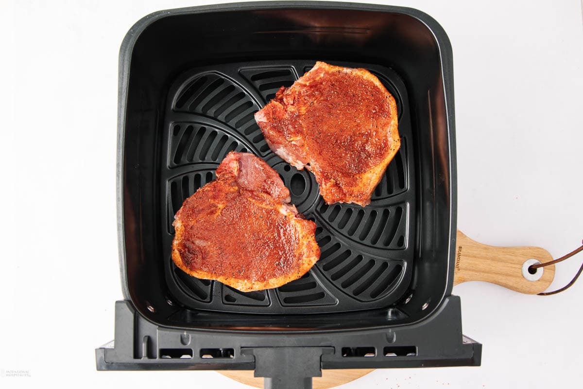 Two seasoned pork chops sit inside the black basket of an air fryer, ready to be cooked. The air fryer rests on a white surface, and the wooden handle is visible on the right side.