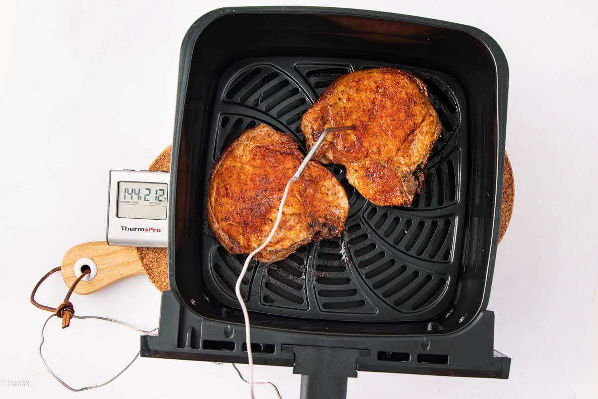 Two cooked pork chops in an air fryer basket with a meat thermometer inserted, displaying a temperature of 142.1°F on its digital screen, all set on a white background.