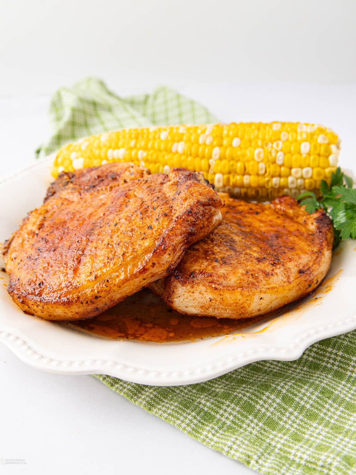 Two seasoned grilled pork chops on a white plate, garnished with sauce, served with a corn on the cob and a sprig of parsley, placed on a green checkered cloth.