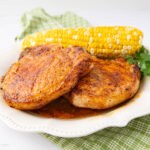 Two seasoned, cooked pork chops on a white plate with a piece of corn on the cob and a sprig of parsley, placed on a green checkered cloth.