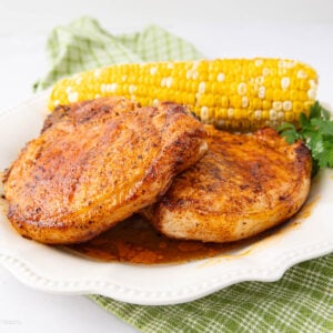 Two seasoned, cooked pork chops on a white plate with a piece of corn on the cob and a sprig of parsley, placed on a green checkered cloth.
