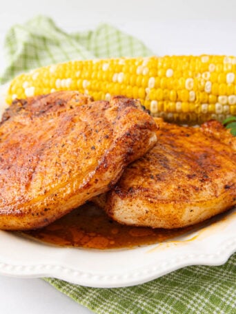 Two seasoned, cooked pork chops on a white plate with a piece of corn on the cob and a sprig of parsley, placed on a green checkered cloth.