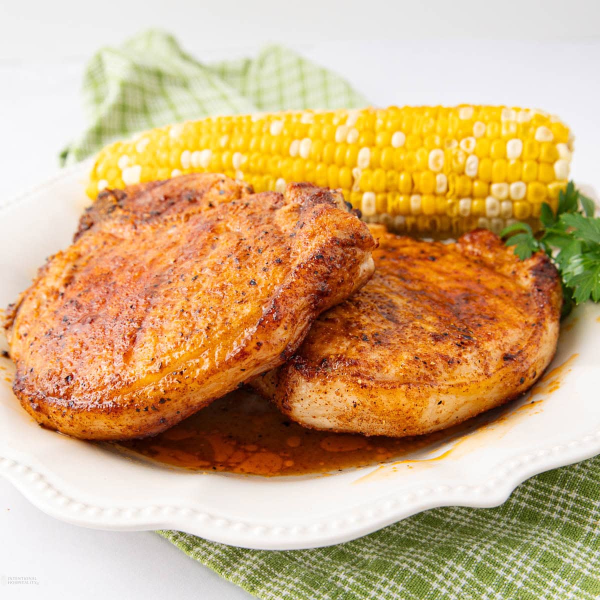 Two seasoned, cooked pork chops on a white plate with a piece of corn on the cob and a sprig of parsley, placed on a green checkered cloth.