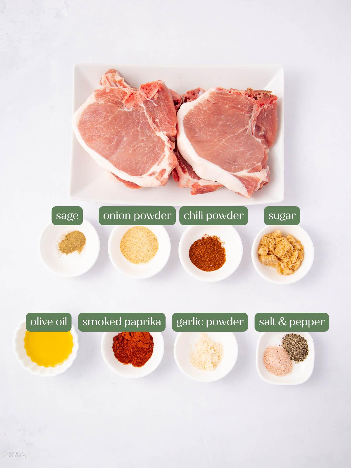 Two raw pork chops on a white plate, with small bowls below containing sage, onion powder, chili powder, sugar, olive oil, smoked paprika, garlic powder, salt, and pepper, all labeled.