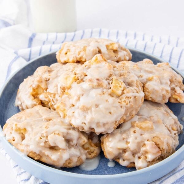 A blue plate stacked with glazed cookies, each filled with chunks of what appears to be apple, resting on a striped cloth with a glass of milk in the background.