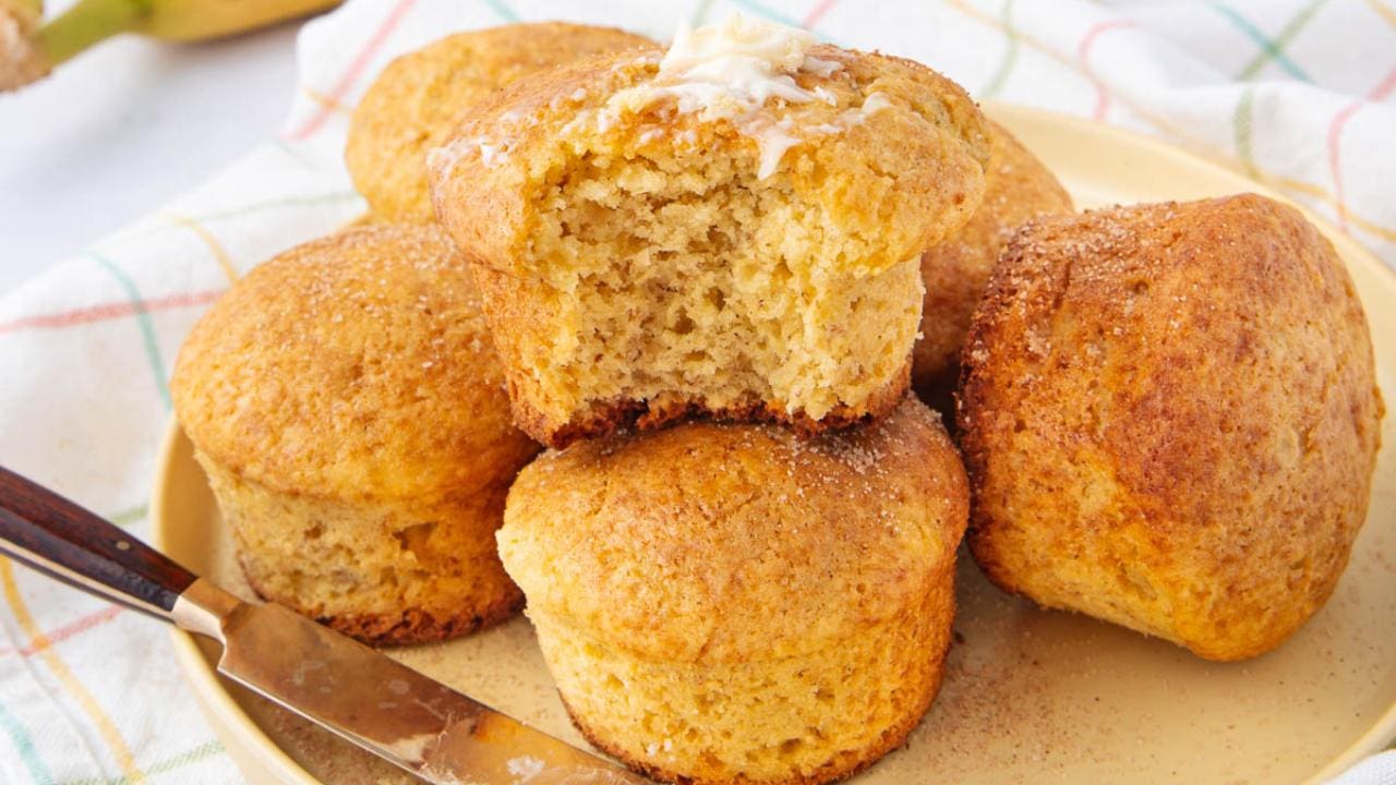 A plate of golden-brown muffins, one with a bite taken out and topped with melting butter. A butter knife rests on the plate, and a checkered cloth is in the background.