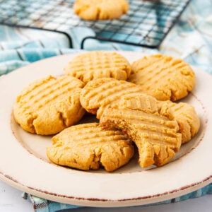 A plate of six peanut butter cookies, five whole and one with a bite taken out, sits on a white plate. A cooling rack with more cookies is visible in the background.