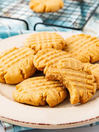 A plate of six peanut butter cookies, five whole and one with a bite taken out, sits on a white plate. A cooling rack with more cookies is visible in the background.