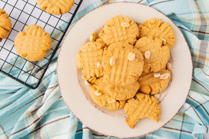 A plate of homemade peanut butter cookies topped with peanuts sits on a blue and white plaid cloth. A cooling rack with more cookies is visible in the corner. The cookies have a crisscross fork pattern on top.