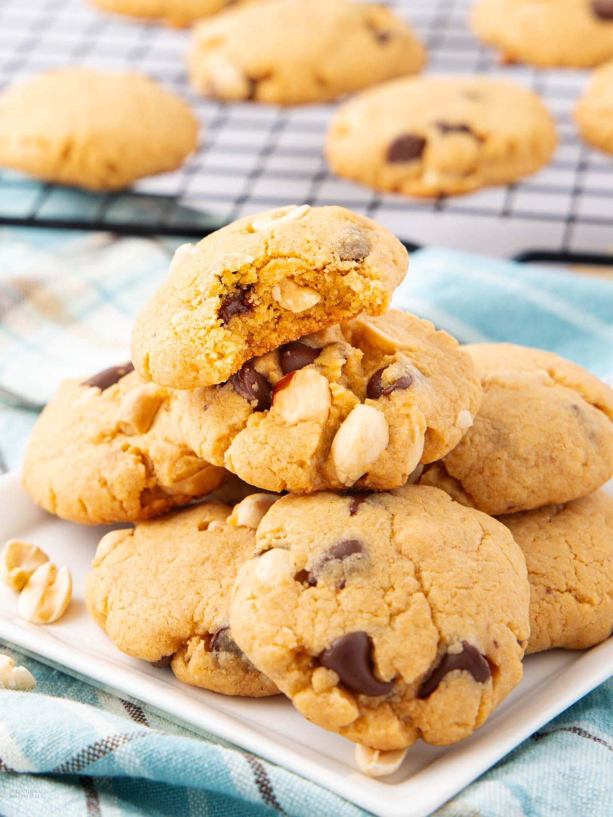A plate of chunky cookies with chocolate chips and nuts, with one cookie on top showing a bite taken out. More cookies are cooling on a wire rack in the background. The scene is set on a blue and white cloth.